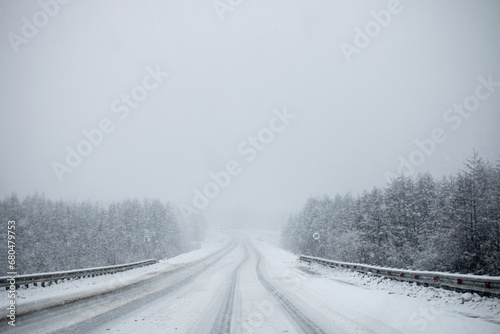 Photo of winter road during the snowfall in Magadan, Russia. Snow showers on trees and hills. Fog and haze, low visibility due to snowstorm. Extreme weather conditions. Car approaching in the distance