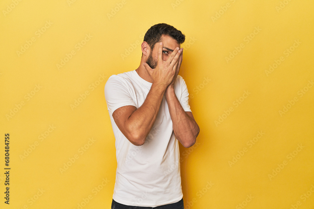 Young Hispanic man on yellow background blink through fingers frightened and nervous.