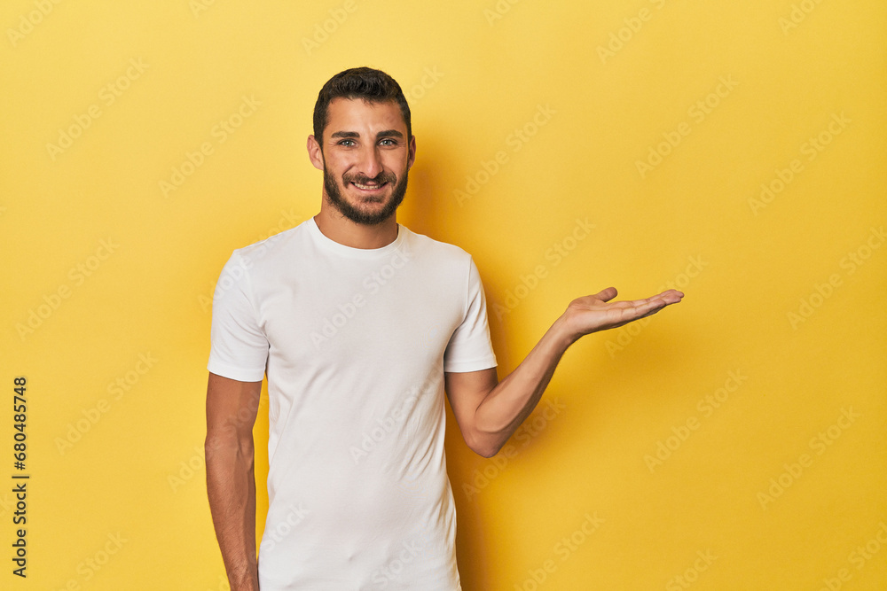 Young Hispanic man on yellow background showing a copy space on a palm and holding another hand on waist. Young Hispanic man on yellow background showing a copy space on a palm and holding another hand on waist.