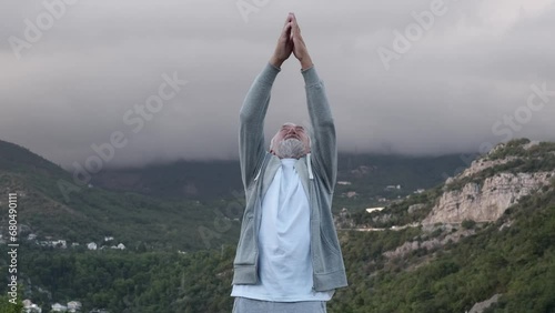 An elderly, gray-haired man with a gray beard in a gray tracksuit practicing yoga against the backdrop of beautiful mountains