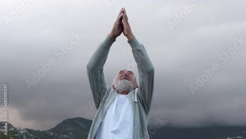An elderly, gray-haired man with a gray beard in a gray tracksuit practicing yoga against the backdrop of beautiful mountains