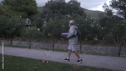 An elderly, gray-haired man with a gray beard in a gray tracksuit holds a yoga mat and sports equipment, against the backdrop of beautiful mountains.