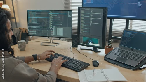 Medium arc shot of young man with long hair and beard, in sweatshirt sitting at desk at home or in office, and typing on keyboard, while writing computer program in coding software on large screen photo