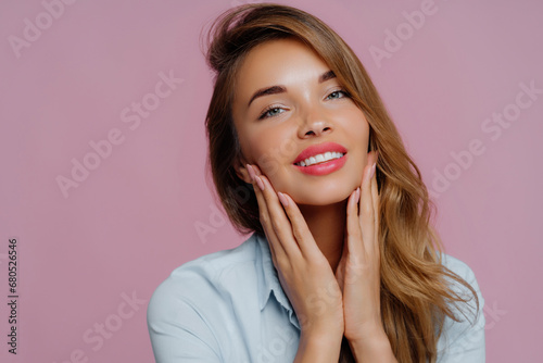 Glowing young woman with hands on face, showcasing flawless skin, smiling on a pink background