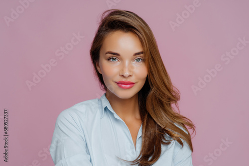 Stunning woman with flowing hair and blue shirt, offering a soft smile against a pink background