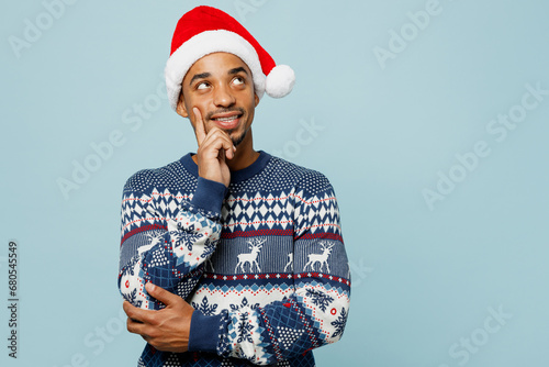 Young man wear knitted sweater Santa hat posing put hand prop up on chin, lost in thought and conjectures isolated on plain blue background. Happy New Year 2024 celebration Christmas holiday concept.