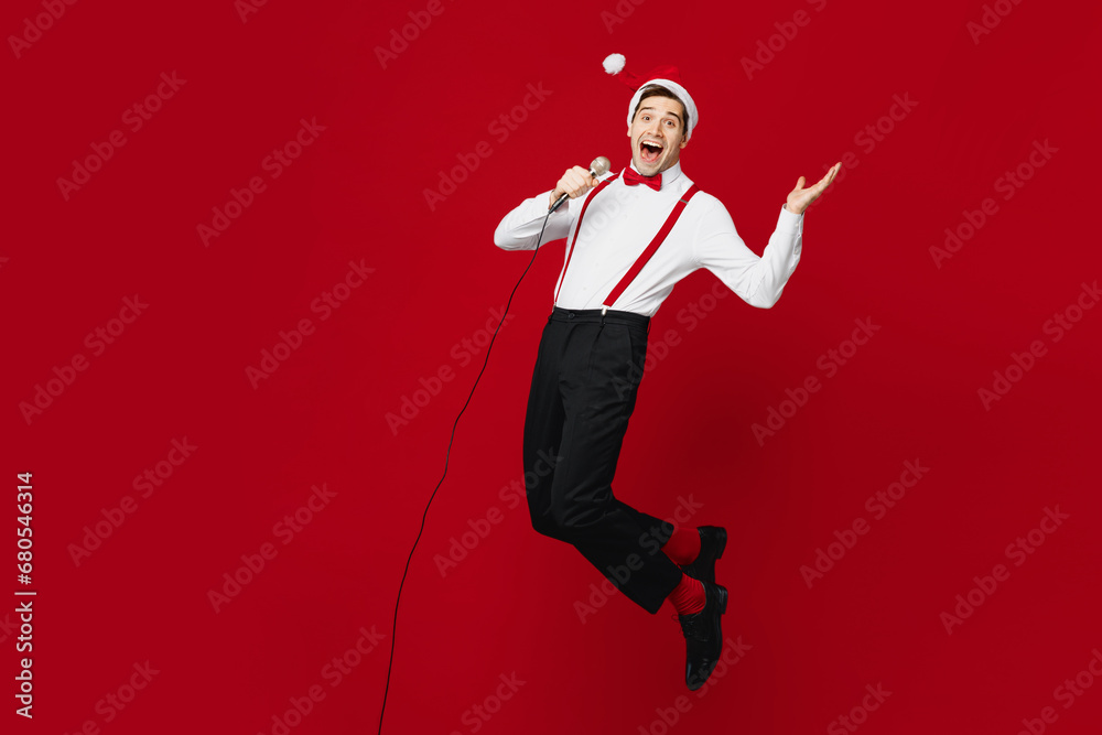Full body side view merry young man wear white shirt Santa hat posing jump high sing song in microphone isolated on plain red background studio. Happy New Year Christmas celebration holiday concept.