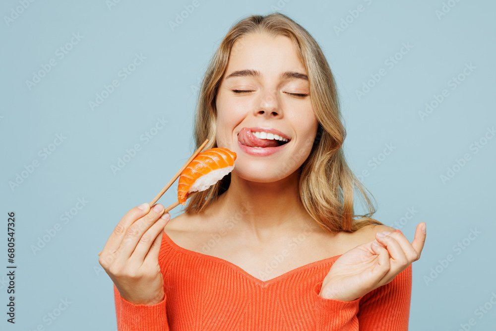 Close up young happy fun woman wear orange casual clothes close eyes hold eat raw fresh sushi roll Japanese food with chopsticks isolated on plain blue background studio portrait. Lifestyle concept. Close up young happy fun woman wear orange casual clothes close eyes hold eat raw fresh sushi roll Japanese food with chopsticks isolated on plain blue background studio portrait. Lifestyle concept.