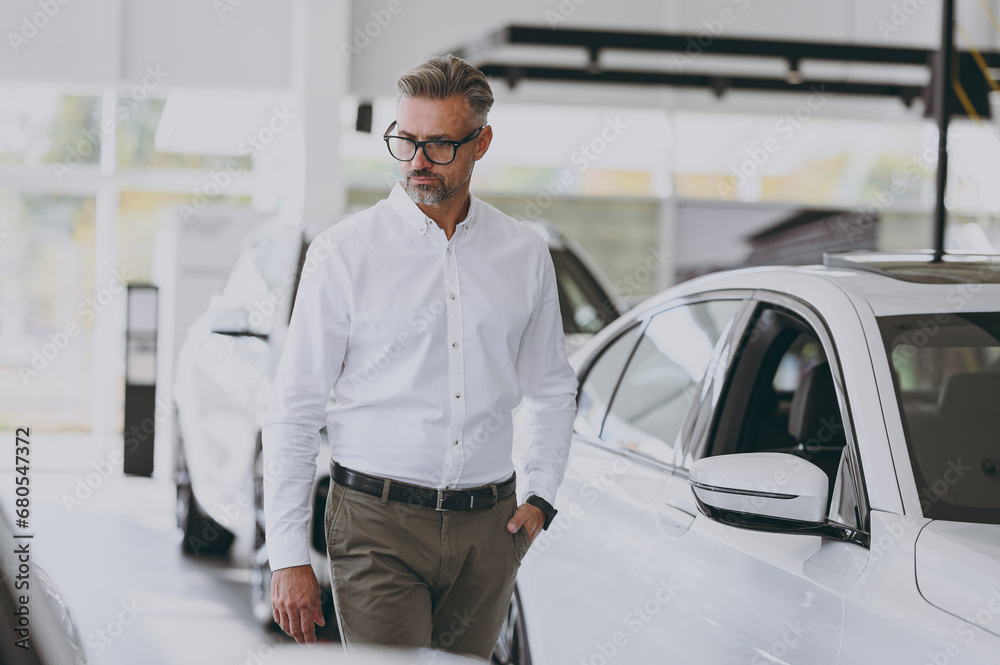 Adult confident calm man customer male buyer client wear classic shirt walk go choose look at auto want to buy new automobile in car showroom vehicle salon dealership store motor show. Sales concept. Adult confident calm man customer male buyer client wear classic shirt walk go choose look at auto want to buy new automobile in car showroom vehicle salon dealership store motor show. Sales concept.