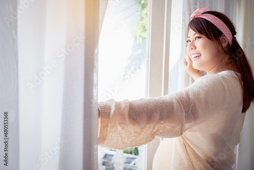 Asian happy pregnant woman standing beside window and open curtain in the morning Pregnancy girl just wake up in the morning and get fresh air Pregnancy motherhood Health care and expectation concept