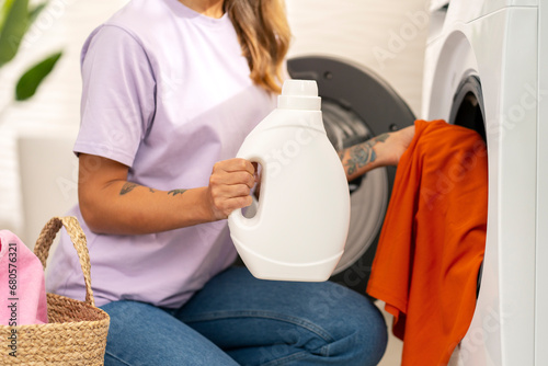 Closeup woman, housekeeper holding bottle with laundry detergent load dirty clothes into washing machine. Mockup. Laundry room concept