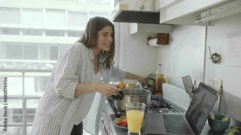Young Hispanic woman cooking food in the kitchen and speaking with senior mother via video call on laptop during the day at home Young Hispanic woman cooking food in the kitchen and speaking with senior mother via video call on laptop during the day at home