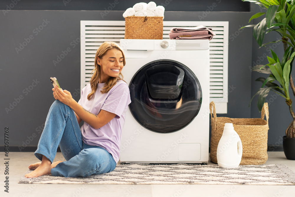 Beautiful smiling woman, housewife holding mobile phone sitting on carpet looking away in modern laundry room with washing machine, laundry detergent, basket with clothes on background. Advertisement - obrazy, fototapety, plakaty 