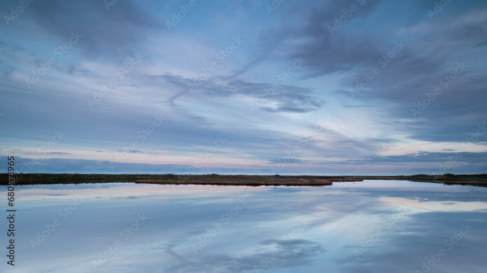 Vast tranquility in the Vatrop nature reserve by the Wadden Sea. The soft clouds are gently colored by the setting sun and reflect in the calm water.