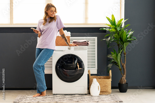 Attractive smiling woman, housekeeper holding mobile phone looking away standing in modern laundry room near washing machine, Advertisement of liquid laundry detergent, mockup