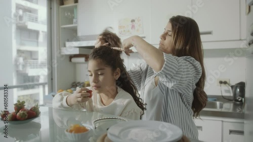Young careful mother tying curly hair of little daughter in a ponytail as girl eating strawberry in the kitchen at home photo