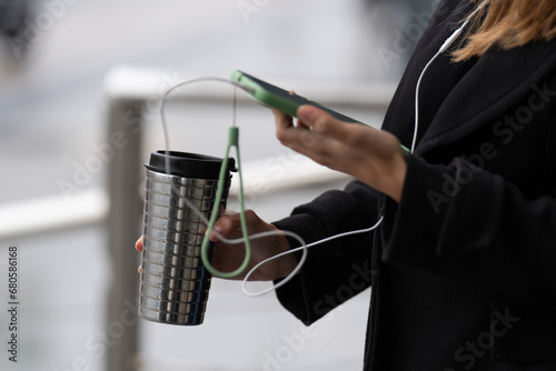 Irrecognizable girl's hand: phone, headphones, steel coffee thermos, multitasking elegance. photo