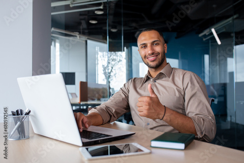 Happy african american businessman sitting in light office at desk, working on laptop, showing thumb up, smiling and looking at camera.