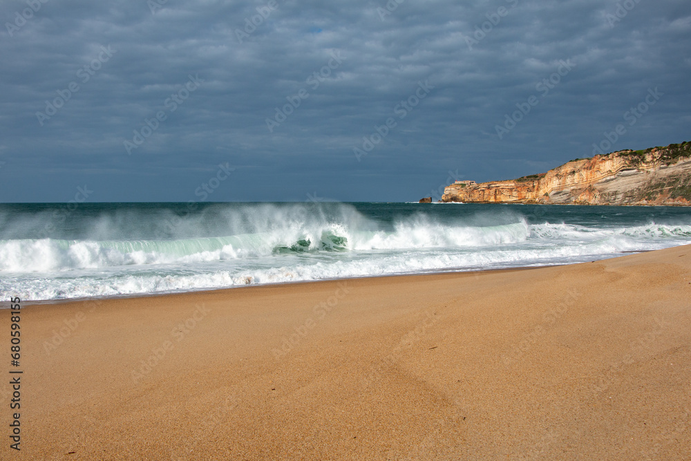 Winter waves breaking on the beach at Nazaré, Portugal, with cliff and lighthouse in the background