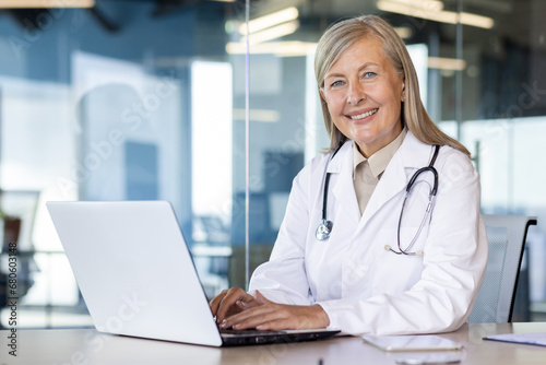 Portrait of mature experienced female doctor, gray-haired senior woman in white medical coat smiling looking at camera, consulting patients remotely, sitting at table with laptop inside clinic.