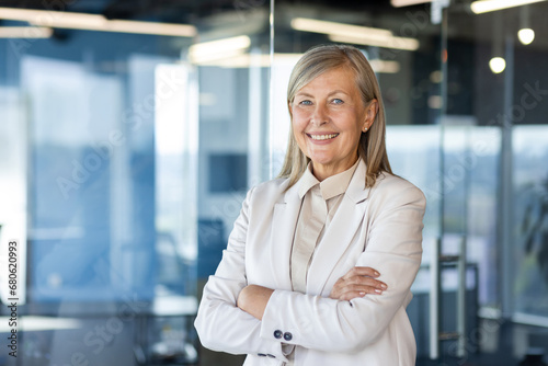 Portrait of mature successful senior woman boss inside office in business suit, businesswoman smiling and looking at camera, standing by window with crossed arms, banker finance woman. photo