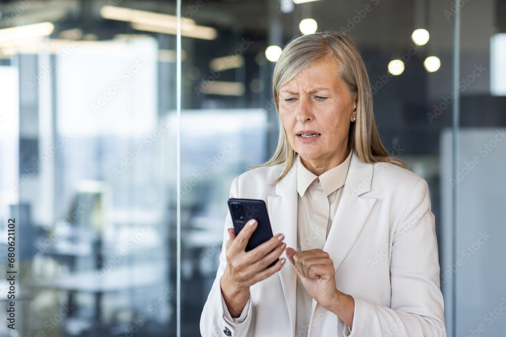 Senior mature woman boss at workplace received online notification message on phone with bad news, financier disappointed sad standing near window inside office, using smartphone. Senior mature woman boss at workplace received online notification message on phone with bad news, financier disappointed sad standing near window inside office, using smartphone.