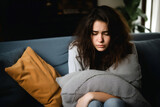 Anxious, nervous oung woman sitting on a couch, embracing a pillow, looking downcast.