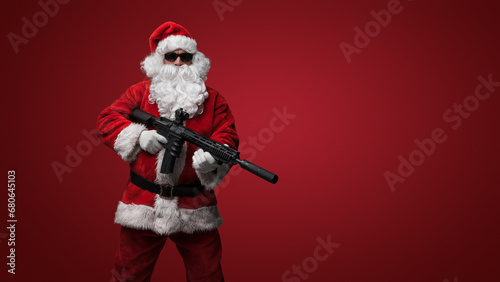 A man dressed as Santa Claus, wearing sleek black sunglasses, poses with toy machine guns against a bold red backdrop