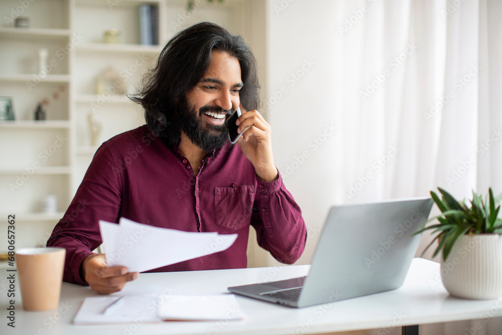 Indian Freelancer Working With Laptop And Papers, Talking On Cellphone At Home Indian Freelancer Working With Laptop And Papers, Talking On Cellphone At Home