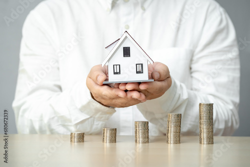 Businessman hand hold a home model on the stacked coin with many increase value on the desk in the office, Financial of real estate business investment and buy house concept.
