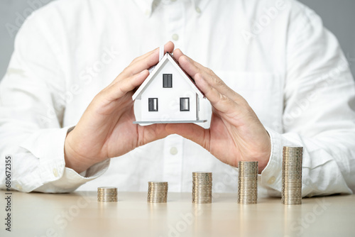 Businessman hand hold a home model on the stacked coin with many increase value on the desk in the office, Financial of real estate business investment and buy house concept.
