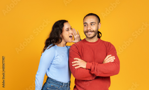 Young woman sharing secret with her husband, yellow background