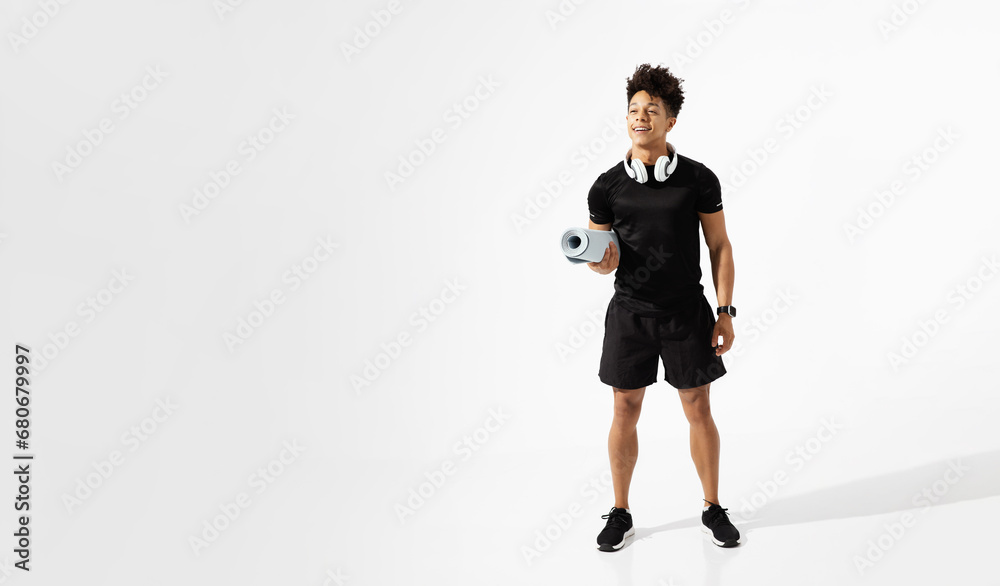 Athletic latin man with headphones holding fitness mat, white background
