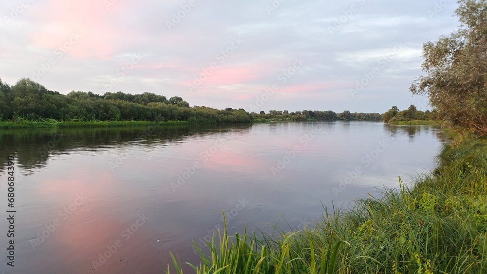 On a quiet summer evening, after sunset, the sky with clouds over the river turns pink and is reflected in the calm water. Shrubs and trees grow on the grassy banks. Branches hang over the water