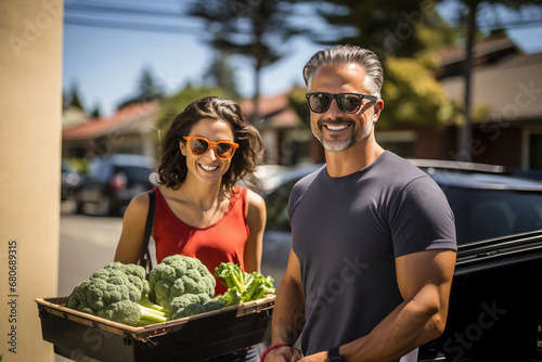 A smiling couple in casual wear and sunglasses carrying fresh vegetables on a sunny suburban street. photo