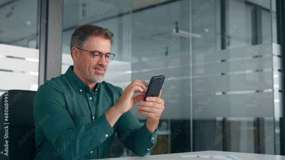 Smiling mid aged businessman executive using cell phone at work desk. Happy busy mature older professional business man manager investor checking finance apps on smartphone in office looking at mobile