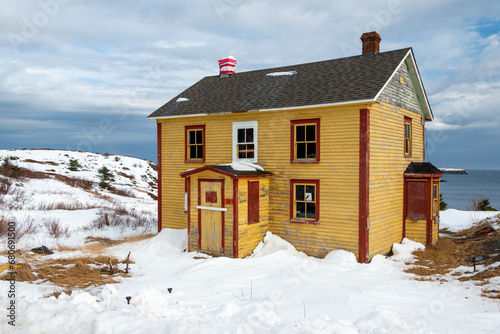 An old yellow wooden two story residence with vibrant red trim, glass windows and a wooden single door. The building is in the middle of a snow covered field with blue ocean and sky in the background.