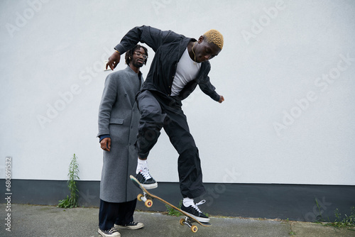 Gen Z black boy wearing stylish jumpsuit shows off his skateboard moves on sidewalk to his friend