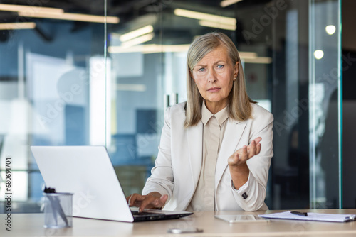 Portrait of a senior gray-haired woman sitting in the office, working on a laptop, spreading her hands in frustration and concern, looking seriously at the camera.