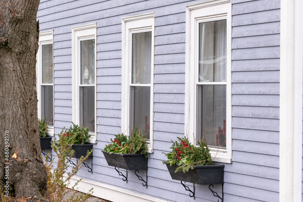 A light blue wooden horizontal clapboard covered house with double hung pane windows. The wooden trim around the window is white. Black flowerboxes hang under the windows and are filled with greenery
