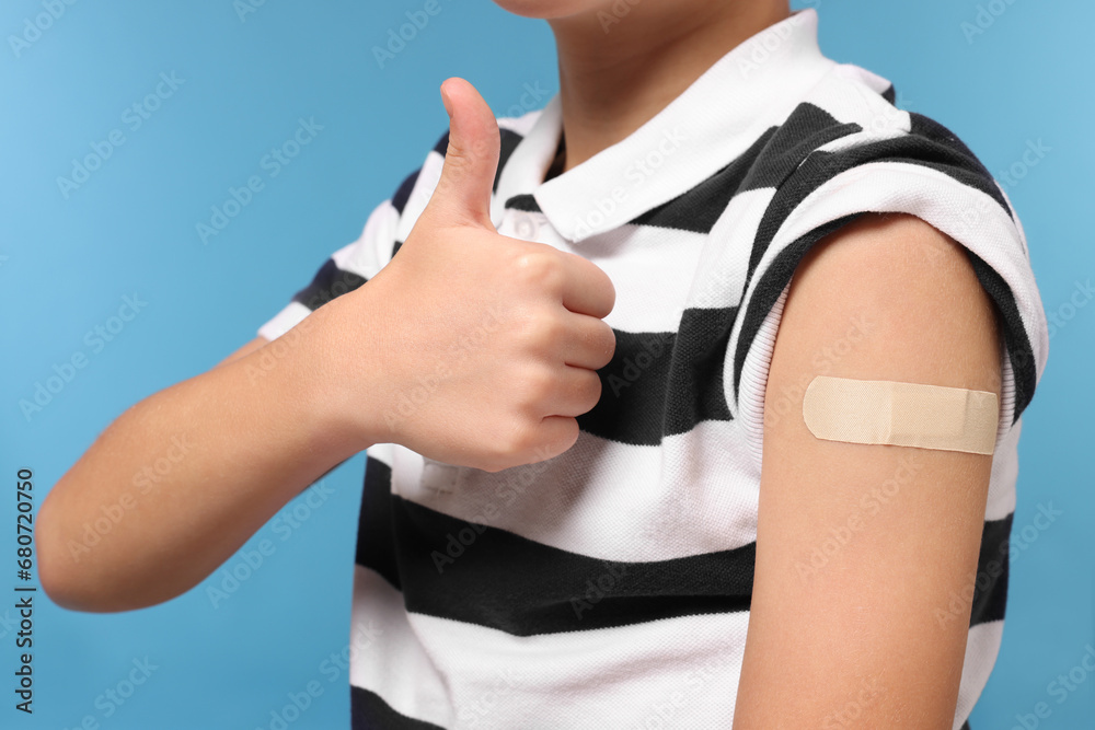 Boy with sticking plaster on arm after vaccination showing thumbs up against light blue background, closeup