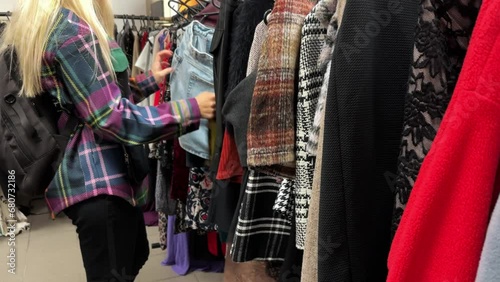 A young woman buys clothes in a second-hand shop. Thrift store hangers with skirts and coats. Fast fashion industry waste reduce and sustainability concept. Selective focus on foreground.