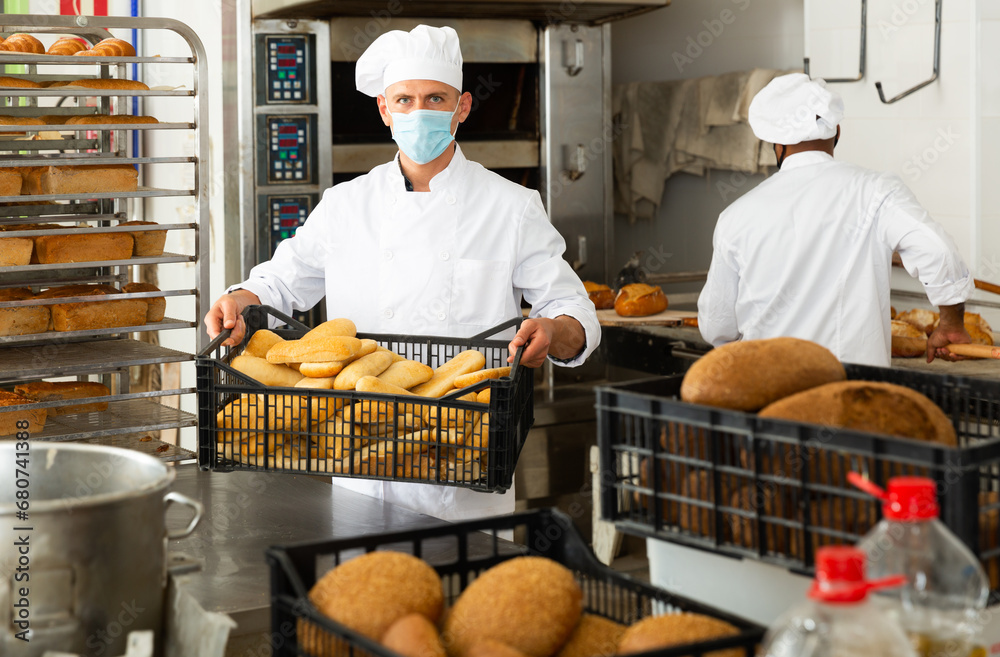 Positive man worker of bakery in protective mask for disease prevention holding box of bread standing at kitchen
