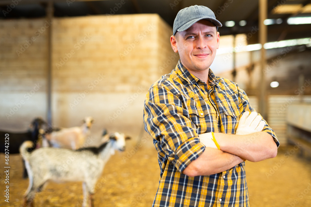 Obraz premium Confident positive male farmer, engaged in breeding dairy goats, posing in goat shed ..