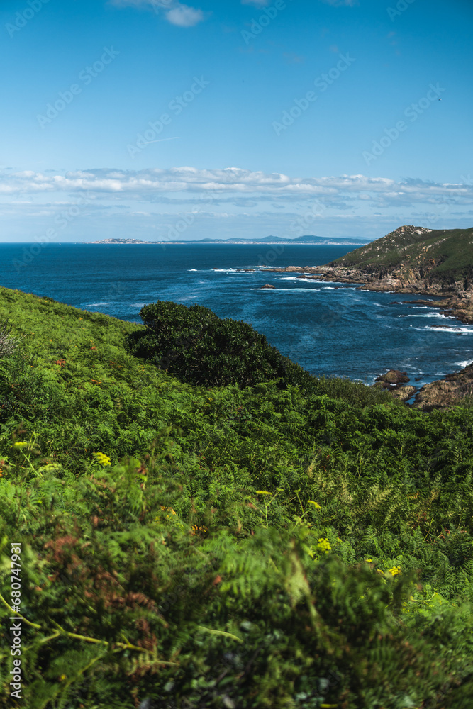 Captivating Ons Island scene: lush greenery in the foreground, azure sea, rocky cliffs, Pontevedra estuary, and a summer sky with scattered clouds from the Buraco do Inferno path.