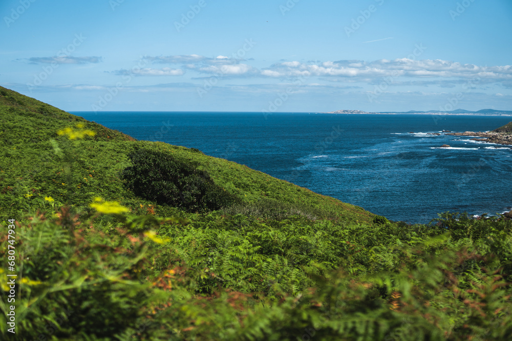 Summer morning view of the Pontevedra estuary from the path to Buraco do Inferno on Ons Island. Lush greenery in the foreground, with the blue Atlantic Ocean beyond under a clear sky.