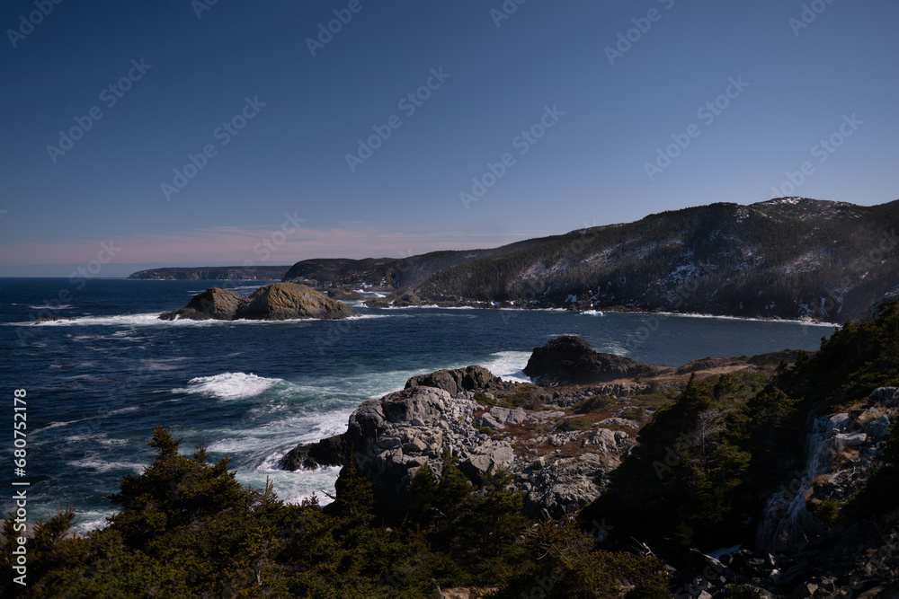 A rugged coastline of rock, cliffs, and boulders along the shoreline in Newfoundland. The sky is deep blue with a pink haze along the horizon of the ocean. The water has waves rolling on shore. 