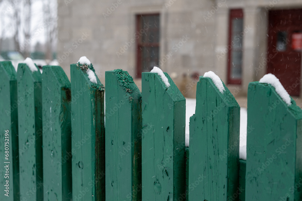 A row of vibrant green color wood fence palings is in the foreground and a large limestone building, trees, and lampposts are in the background. The tall picket fence has been newly painted. 