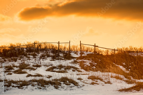 A dramatic orange evening sky with dark clouds over a grassy hill covered with grass, hay and white fresh snow in a meadow. There's an old wooden farm fence on the hillside with a small opening. 