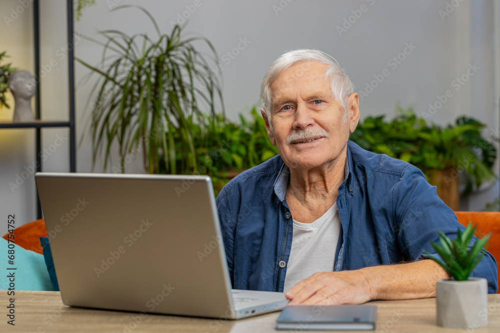 Happy senior man sitting on sofa at table working on laptop pc at office home room. Elderly mature grandfather pensioner freelancer online remote job. E-learning browsing internet on notebook computer Happy senior man sitting on sofa at table working on laptop pc at office home room. Elderly mature grandfather pensioner freelancer online remote job. E-learning browsing internet on notebook computer
